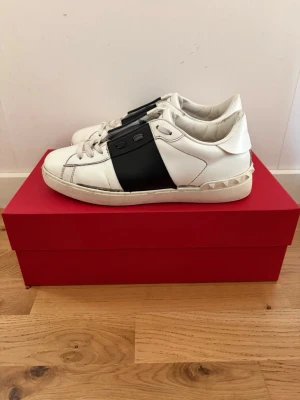 Valentino Garavani Open Sneakers - Snygga vita sneakers från Valentino Garavani med bred svart rem över mitten och ikoniska nitar på hälen. Skorna är i läder och har en klassisk låg profil med vita snören och detaljer. Perfekta för dig som gillar exklusiv streetstyle. Skorna kommer med boxen, dustbags och ett extra par skosnören. Sulan har lossnat lite på båda skorna men det påverkar inte funktionen och går enkelt att fixa med superlim.