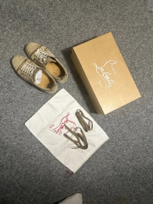 Beige sneakers från Christian Louboutin - Stilrena beige sneakers från Christian Louboutin i mocka med vita snören och klassisk låg siluett. Skorna har diskreta sömdetaljer och kommer med originalkartong, extra skosnören och dustbag. Perfekta för dig som gillar exklusiva och tidlösa sneakers.   