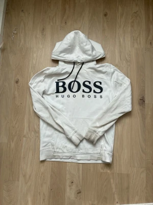 Vit hoodie från Hugo Boss - Snygg vit hoodie från Hugo Boss med stor svart logga på bröstet och svarta dragsnören i huvan. Klassisk känguruficka framtill och mjukt material som känns skönt mot huden. Perfekt för en avslappnad och trendig stil.