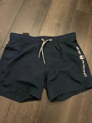 Marinblå badbyxor från Sail Racing - Snygga marinblå badbyxor från Sail Racing med vit snörning i midjan och logga längs ena benet. Klassisk modell med elastisk midja och bakficka. Perfekta för stranden eller poolen.