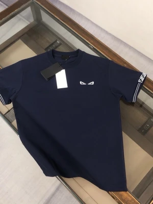 Svart Fendi t-shirt med logga - Snygg svart t-shirt från Fendi med korta ärmar och ikoniska Fendi-loggor på ärmsluten. T-shirten har en liten vit detalj på bröstet som liknar ögon och är tillverkad i mjuk bomull. Perfekt för en trendig och avslappnad stil.