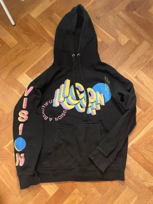 Svart SYRE hoodie med färggrafik - Cool svart hoodie från SYRE med stor färgglad 3D-illusion- och textgrafik på både fram- och baksida. Tryck i gult, rosa, blått och lila, samt texten 'VISION' längs ena armen. 