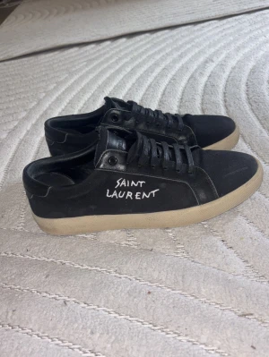 Saint Laurent skor  - Svarta sneakers från Saint Laurent med vit text på sidan. Skorna har låga skaft, svarta skosnören och beige sula. Materialet ser ut att vara en mix av mocka och läder vilket ger en clean och stilren look. 
