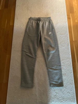Gråa mjukisbyxor från Nike - Gråa mjukisbyxor från Nike med elastisk midja och två sidofickor. Byxorna har en smal passform och är tillverkade i mjukt material med Nike-logga tryckt på vänster lår. Perfekta för chill eller träning.