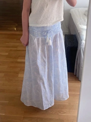Ljusblå maxikjol med smock och tofsar - Luftig ljusblå maxikjol med smock i midjan och knytband med tofsar. Kjolen har ett diskret mönster och är tillverkad i ett tunt, flowy tyg som ger en avslappnad känsla. 