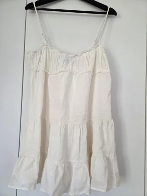 Vit sommarklänning från Zara - Luftig vit klänning från Zara med tunna axelband och volangdetaljer. Klänningen har en avslappnad passform och är tillverkad i ett mjukt bomullstyg, perfekt för varma dagar. Enkel och stilren design som ger en fräsch look.