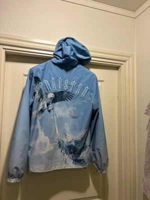 Blå vindjacka från Trapstar med tryck - Snygg blå vindjacka från Trapstar med huva och elastiska muddar. Nypris på butik var 2500kr 