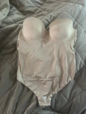 Beige body med vadderade kupor - Snygg beige body med vadderade kupor och axelbandslös design. Tillverkad i ett stretchigt och mjukt material som formar sig efter kroppen. Perfekt för att skapa en slät siluett under kläder. Knäppning i grenen för enkel användning.