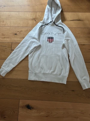 Vit hoodie från GANT med logga - Snygg vit hoodie från GANT med broderad logga och sköld på bröstet. Klassisk känguruficka framtill och svart dragsko i huvan. Perfekt för en avslappnad och stilren look.