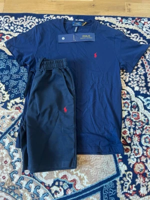 Mörkblå set från Polo Ralph Lauren - Snyggt mörkblått set från Polo Ralph Lauren med t-shirt och shorts. Båda plaggen har det klassiska röda Polo-logot broderat på bröstet och benet. Setet är gjort i mjuk bomull och har en avslappnad passform med elastisk midja på shortsen.