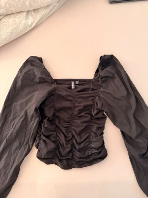 Svart topp med puffärmar från Pieces - Trendig svart topp från Pieces med långa puffärmar och rynkade detaljer framtill. Fyrkantig urringning ger en snygg siluett och toppen är tillverkad i ett mjukt tyg som sitter bekvämt. Perfekt för dig som gillar en stilren men ändå cool look. Stolek XS, använd 1-2 gånger