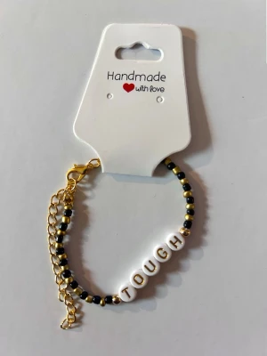 Pärlarmband med texten TOUGH - Handgjort armband med svarta och guldiga pärlor samt bokstavspärlor som bildar ordet TOUGH. Armbandet har guldfärgad kedja och lås, perfekt för att ge en personlig touch till din stil.