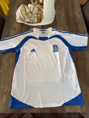 Grekland Adidas fotbollströja - Vit och blå fotbollströja från Adidas med Greklands landslagsemblem på bröstet. Tröjan har klassiska Adidas-ränder på axlarna och är tillverkad i ett lätt och ventilerande material, perfekt för träning eller match.