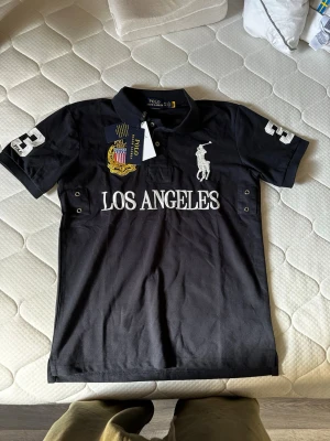 Mörk blå  pikétröja Polo Ralph Lauren - Svart pikétröja från Polo Ralph Lauren med vit broderad logga, texten 'LOS ANGELES' och siffran 3 på både ärm och rygg. Klassisk krage och korta ärmar, tillverkad i bomull. Snygga detaljer med broderad patch och knappar vid sidorna.
