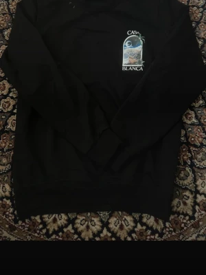 Casablanca tröja - Säljer en svart sweatshirt från Casablanca med tryck både fram och bak. På bröstet och ryggen finns ett färgglatt motiv med stadsvy, palmer och texten 'Casa Blanca'. Tröjan har rund hals och ribbade muddar. Perfekt för dig som gillar streetwear och snygga prints. Inga slitelser och väl omhändetagen