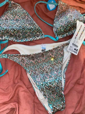 Mönstrad bikini med turkosa detaljer - Den har fått en smink fläck på underdelen, men har ej använts, Säljer en snygg bikini från Calzedonia med färgglatt mönster i turkos, rosa, svart och vitt. Överdelen har trekantsform och knytband i turkost, underdelen har samma mönster och smala sidor. Perfekt för stranden eller poolen!