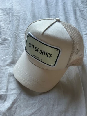 Beige keps John Hatter & Co - Snygg beige keps från John Hatter & Co med mesh på sidorna och bak. Framtill sitter en metallskylt med texten 'OUT OF OFFICE' som ger en cool och avslappnad vibe. Perfekt accessoar för en trendig streetstyle-look.