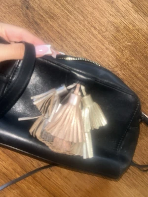 Svart handväska från Rebecca Minkoff - Pris kan diskuteras!!!!! !!!!  Snygg svart väska i läder från Rebecca Minkoff med dekorativa tofsar i metallicfärger. Väskan har en stilren design med dragkedja och en liten metallogo framtill. Perfekt för dig som gillar trendiga accessoarer. 