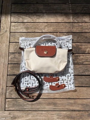 Beige Longchamp Le Pliage handväska - Snygg beige handväska från Longchamp Le Pliage med bruna detaljer i läder och guldfärgad knapp. Väskan har ett klassiskt, stilrent utseende och levereras med axelrem och extra läderdetaljer. Perfekt för dig som gillar tidlös design och praktiska accessoarer.