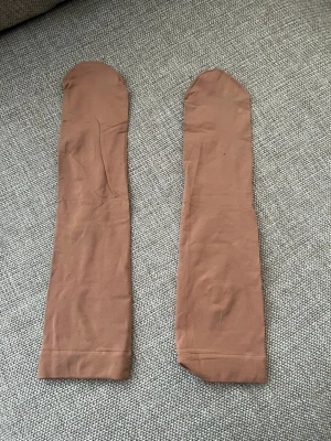 Beige tunna nylonstrumpor - Säljer ett par tunna, beige nylonstrumpor med klassisk design. Perfekta att bära under kjol eller klänning för en naturlig look. Strumporna är långa och har en mjuk, elastisk kant upptill för bekväm passform.