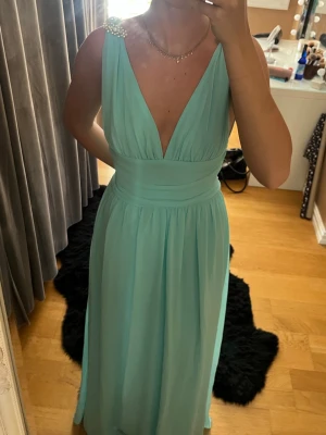 Ljusblå långklänning med pärldetaljer - Elegant ljusblå långklänning med djup v-ringning och bred midja. Klänningen har pärldetaljer på axlarna och är ärmlös. Perfekt för speciella tillfällen och har ett luftigt, svepande fall.