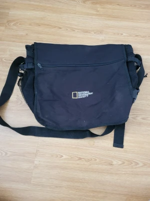 National Geographic Society durable bag  - Weather resistant and durable fabric. This bag features an adjustable buckle for the main pocket, two large adjustable side pockets for waterbottles etc, one window pocket, 3 pen pockets, one mesh zipped pocket that unfortunately has some tearing, one clip, one standard smaller pocket inside, and large zippered pocket in the front. Velcros shut and adjustable strap with very durable clip. Signs of use. Everything works perfectly, but mesh pocket has tearing. See other ads for boundle discounts.