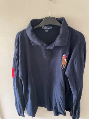 Mörkblå långärmad pikétröja Polo Ralph Lauren - Mörkblå långärmad pikétröja från Polo Ralph Lauren med klassisk krage och broderad polospelare på bröstet. Röd detalj på ena ärmen. Tillverkad i mjuk bomull, perfekt för en avslappnad men stilren look.