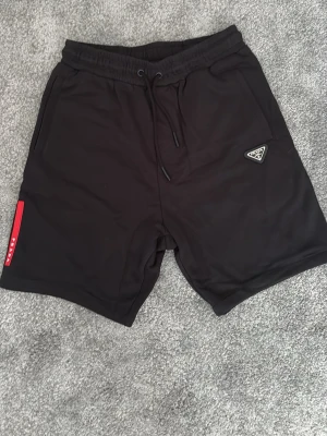 Svarta shorts från Prada med snörning - Svarta shorts från Prada med elastisk midja och snörning. Klassisk design med två fickor fram och en bakficka. Diskret Prada-logga på vänster ben och röd detalj på sidan. Perfekta för en stilren och avslappnad look.