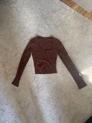 Brun spetsig långärmad topp - Snygg brun långärmad topp i spetsmaterial med knytning framtill och djup urringning. Figurnära passform och croppad längd, perfekt att styla med jeans eller kjol. Tunn och stretchig känsla.