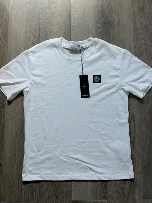 Vit Stone Island t-shirt - Säljer en klassisk vit t-shirt från Stone Island med rund hals och korta ärmar. T-shirten har en svart patch med logga på bröstet och är tillverkad i mjuk bomull. Perfekt basplagg för en clean och stilren look.