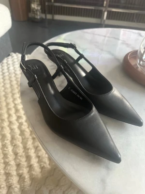 Svarta slingback pumps med smal klack - Stilrena svarta pumps från XII med spetsig tå och smal slingback-rem. Skorna har en elegant låg klack och är tillverkade i ett slätt material som ger en klassisk look. Perfekta för dig som gillar minimalistisk och trendig stil.