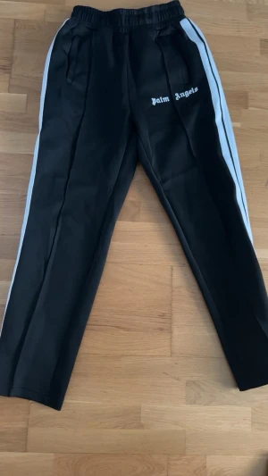Palm Angels svarta mjukisbyxor - Svarta mjukisbyxor från Palm Angels med vita sidoränder och dragkedja vid benslutet. Byxorna har resår i midjan, fickor och logga broderad på vänster lår. Snygg streetwear-look och sköna att bära till vardags.