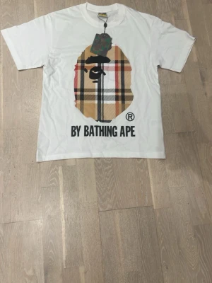 Vit BAPE t-shirt med rutig logga - Vit t-shirt från A Bathing Ape med stor rutig BAPE-logga i gult, svart, rött och vitt på bröstet. Klassisk rund hals och korta ärmar. Tryckt text 'BY BATHING APE' nedanför loggan. Perfekt för dig som gillar streetwear och unika prints.