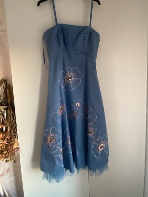 Vintage blå klänning med blommor och paljetter - Söt blå klänning med stora broderade blommor och paljett-detaljer. Klänningen har en lätt utsvängd form och volangkant nedtill. Perfekt för dig som vill sticka ut med en unik och färgglad look. Jättefin till bröllop, bal. Den är inte ful längd man ser anklarna på mig när jag har på mig den (är 167 cm).Banden på klänningen är avtagbara. Min mamma hade den för många årsen❣️