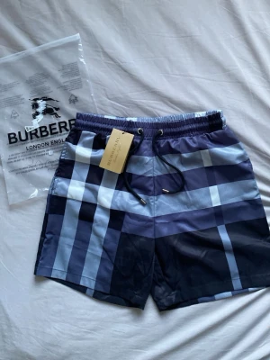 Burberry rutiga badbyxor blå - Snygga badbyxor från Burberry med klassiskt rutigt mönster i olika nyanser av blått och vitt. Modellen har elastisk midja med dragsko och en bakficka. Perfekta för stranden eller poolen, och tillverkade i snabbtorkande material.