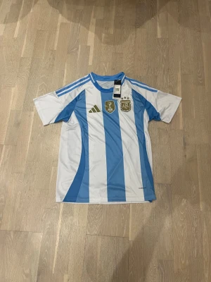 Argentina matchtröja Adidas blåvit - Säljer en officiell Argentina matchtröja från Adidas i klassiska blå och vita ränder. Tröjan har guldiga detaljer, FIFA-märke och AFA-logga på bröstet samt tre blå ränder på axlarna. Tillverkad i lätt och ventilerande material, perfekt för fotboll.