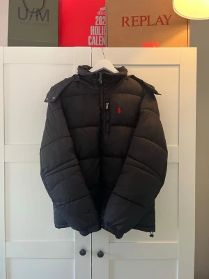 Svart pufferjacka från Ralph Lauren - Svart pufferjacka från Ralph Lauren med huva och dragkedja framtill. Jackan har en klassisk quiltad design och den ikoniska röda loggan på bröstet. Perfekt för kalla dagar och enkel att matcha med olika outfits.