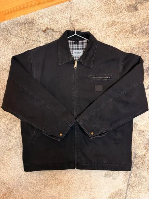 Svart Carhartt jacka med rutigt foder - Svart jacka från Carhartt med dragkedja, klassisk krage och rutigt innerfoder. Jackan har två sidofickor och en bröstficka med dragkedja. Perfekt för dig som gillar en stilren och tidlös look.