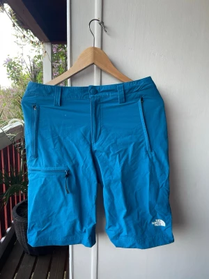 Blå shorts från The North Face - Snygga blå shorts från The North Face med dragkedjeförsedda fickor fram och bak. Shortsen har bälteshällor och är tillverkade i ett lätt och slitstarkt syntetmaterial, perfekta för aktiva dagar utomhus.