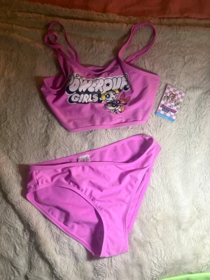 Rosa Powerpuff Pinglorna bikini - Säljer en söt rosa bikini med Powerpuff Pinglorna-tryck på överdelen. Bikinin har sportig topp med smala axelband och matchande trosor. Perfekt för dig som älskar tecknade serier och vill sticka ut på stranden! Den är dessutom oanvänd