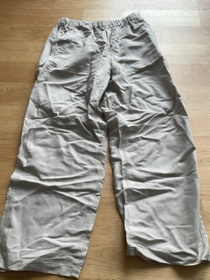 Beiga linnebyxor med raka ben - Säljer ett par beiga linnebyxor med avslappnad passform och raka, vida ben. Byxorna är tillverkade i ett svalt och lätt linnematerial, perfekta för varma dagar. Enkel design utan synliga detaljer eller fickor framtill.