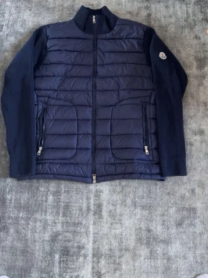 Mörkblå cardiganjacka från Moncler - Snygg mörkblå cardiganjacka från Moncler med quiltad framsida och stickade ärmar. Jackan har två dragkedjefickor framtill, ribbad krage och ärmslut samt Moncler-logga på ena ärmen. Perfekt för lager-på-lager och stilren look.
