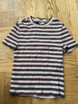 Randig ribbad t-shirt från H&M Divided - Randig t-shirt från H&M Divided i rosa, svart och vit. Modellen är ribbad med korta ärmar och rund halsringning. Perfekt för en avslappnad och trendig look.