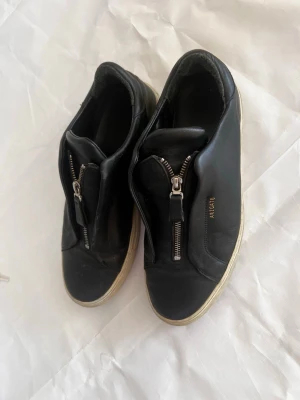 Svarta sneakers från ARIGATO med dragkedja - Svarta sneakers i läder från ARIGATO med cool dragkedja framtill istället för klassiska snören. Vit sula och diskret guldfärgad logga på sidan. Snygg minimalistisk design som passar till det mesta.