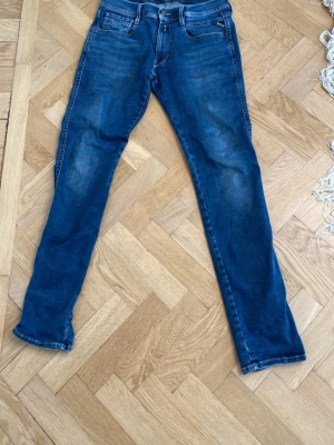 Blå  jeans från Replay - Snygga blåa jeans från Replay med klassisk femficksdesign och diskreta slitningar. Jeansen har normal midja, dragkedjegylf och logotypdetaljer på fickorna. Perfekta för en trendig och avslappnad stil.DEN HAR NÅGRA HÅL! 