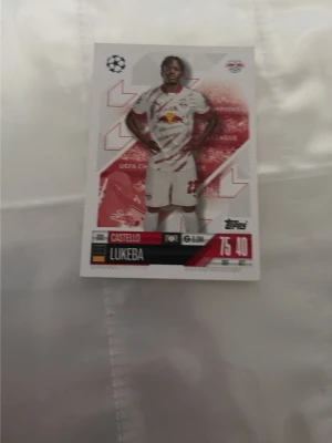 Fotbollssamlarkort RB Leipzig Lukeba - Säljer ett fotbollssamlarkort från Topps med spelaren Castello Lukeba i RB Leipzigs vita matchställ. Kortet har röda och vita detaljer samt spelarens namn och statistik tryckt på framsidan. Perfekt för samlare av fotbollskort.
