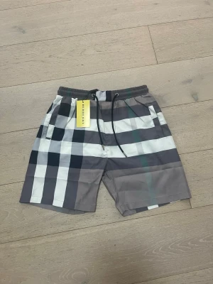 Rutig shorts från Burberry - Snygga shorts från Burberry med klassiskt rutigt mönster i grått, vitt, svart och grönt. Modellen har elastisk midja med snörning och två sidofickor. Perfekta för en trendig och avslappnad stil.