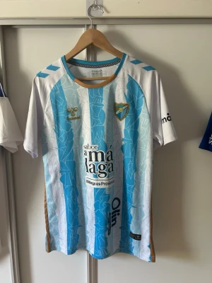 Málaga CF matchtröja blåvit - Snygg fotbollströja från Málaga CF i blått och vitt med guldiga detaljer. Tröjan har klubbmärke på bröstet, sponsortryck och ett unikt mönster över hela tyget. Korta ärmar och rund halsringning. Perfekt för dig som älskar fotboll och vill sticka ut på planen.