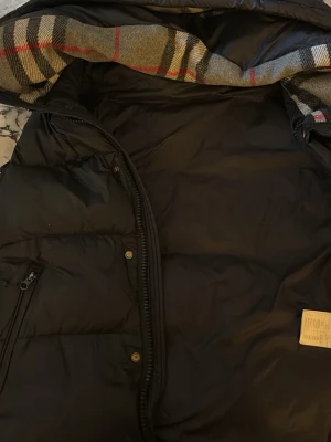 Svart dunjacka med rutigt foder - Snygg svart dunjacka med dragkedja och tryckknappar. Insidan av huvan har ett klassiskt rutigt foder i grått, svart, beige och rött. Jackan har en ficka med dragkedja på bröstet och är perfekt för kalla vinterdagar.