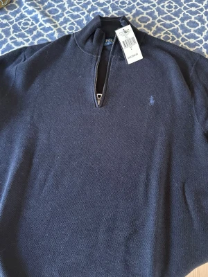 Mörkblå halvzip från Polo Ralph Lauren - Mörkblå långärmad tröja från Polo Ralph Lauren med halvzip och broderad logga på bröstet. Klassisk och stilren modell med hög krage. Perfekt för lager-på-lager och snygg till jeans eller chinos.